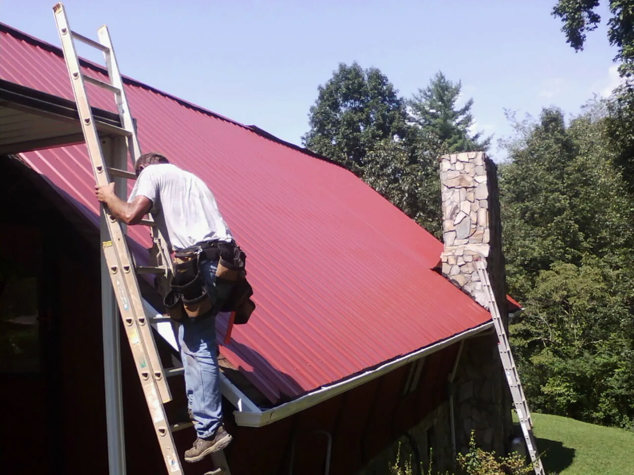 Roofer on a ladder beside a red metal roof and stone chimney — Soffit & Fascia Repair by Roofing Brothers in Minnetonka